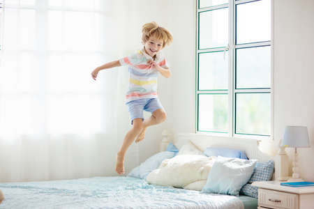 Child jumping on bed in white bedroom with big window. Kids play at home. Fun jump on parents bed. Room and interior design. Boy at home. Sunny morning, weekend family relaxing time.の写真素材