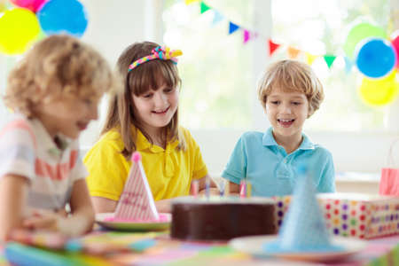 Kids birthday party. Child blowing candles on cake and opening presents. Pastel rainbow theme celebration. Family celebrating at home. Boy opening gifts, eating cakes. Sweets for children.の写真素材