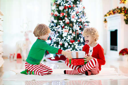 Children at Christmas tree and fireplace on Xmas eve. Family with kids celebrating Christmas at home. Boy and girl in matching pajamas decorating xmas tree and opening presents. Holiday gifts for kid.の写真素材