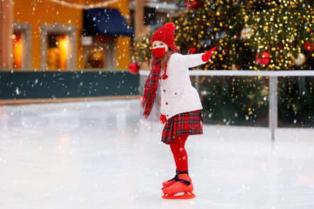Kid in face mask ice skating in winter park rink. Safe Christmas fun in covid-19 pandemic. Coronavirus prevention in holiday season. Children ice skate on Xmas fair. Little girl with skates.の写真素材