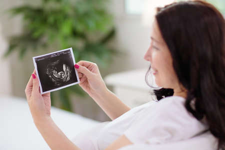 Pregnant woman at home. Young expecting mother in white bedroom. Healthy pregnancy. Female looking at baby sonogram preparing for birth. Maternity and prenatal care.の写真素材