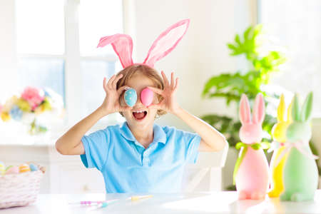 Kids dyeing Easter eggs. Children in bunny ears dye colorful egg for Easter hunt. Home decoration with flowers, basket and rabbit for spring holiday celebration. Little boy decorating home.の写真素材