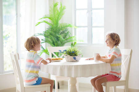 Kids eating breakfast in white kitchen. Children eat sandwich and drink juice on sunny morning. family at home. Boy cooking lunch. House dining room interior. child healthy nutrition.の写真素材