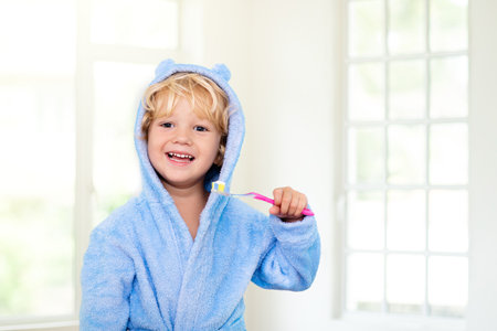 Child brushing teeth. Kids toothbrush and paste. Little baby boy in blue bath robe or towel brushing his teeth in white bathroom with window on sunny morning. Dental hygiene and health for children.の写真素材