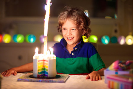 Kids birthday party. Child blowing candles on cake and opening presents. Pastel rainbow theme celebration. Family celebrating at home. Boy opening gifts, eating cakes. Sweets for children.の写真素材