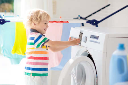 Child in laundry room with washing machine or tumble dryer. Kid helping with family chores. Modern household devices and washing detergent in white sunny home. Clean washed clothes on drying rack.の写真素材