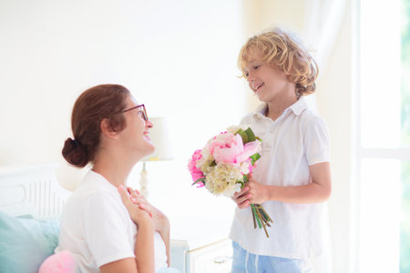 Child giving mom present in bed. Mother's Day morning gift. Little boy with flower bouquet hugging his mom in white sunny bedroom. birthday celebration. Breakfast in bed on mothers day. family love.の写真素材