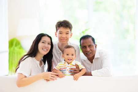 Happy Asian family with kids at home. Parents and children sitting on white couch in sunny living room. Mother, father, son and daughter play and laugh. Mature interracial couple in new house.の写真素材