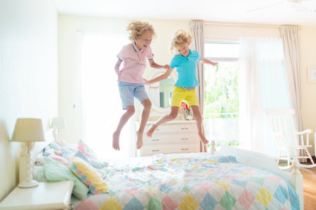 Child jumping on bed in white bedroom with big window. Kids play at home. Fun jump on parents bed. Room and interior design. Boy at home. Sunny morning, weekend family relaxing time.の写真素材