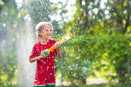 Kids play with water sprinkle hose. Summer garden outdoor fun for children. Boy splashing water on hot sunny day. Kid watering plants in backyard. Toy water gun.の写真素材