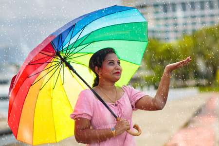 Young woman walking under heavy rain with rainbow umbrella. Indian female having fun in autumn...