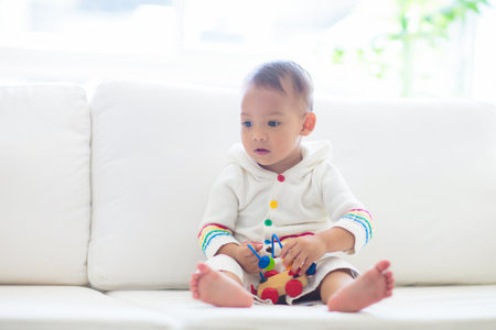 Asian baby boy playing with colorful toy train in white sunny bedroom. Baby with educational toys....