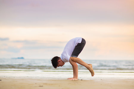 Teenager doing calisthenics exercise. Beach yoga at sunset. Teen boy exercising. Crow and peacock...