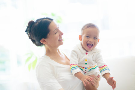Mother and child on a white couch. Asian mom and baby boy in diaper playing in sunny bedroom. Parent and little kid relaxing at home. Family having fun together. Bedding and textile for infant nursery.の写真素材