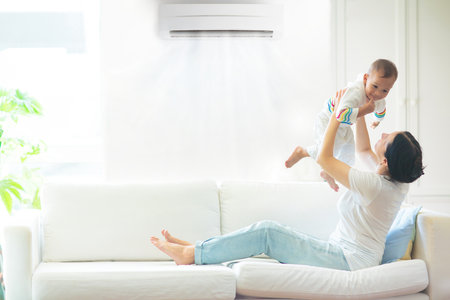 Asian mother and child with air conditioner remote control. Comfortable temperature at family home. Cooling device. Mom and baby boy on couch under cold breeze. Air conditioning on hot summer day.の写真素材