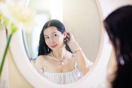 Young Asian woman looking in the mirror. Skin care routine. Beautiful female in a night gown applying cream on date night or weekend morning. Skin cleansing and treatment. Jewelry and pearls.の写真素材