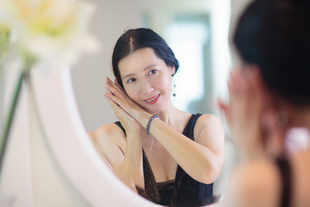 Young Asian woman looking in the mirror. Skin care routine. Beautiful female in a night gown applying cream on date night or weekend morning. Skin cleansing and treatment. Jewelry and pearls.の写真素材