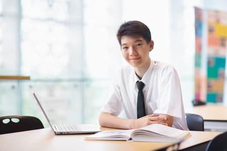Asian boy with laptop in school. Student in classroom. Teenager in uniform doing homework with...