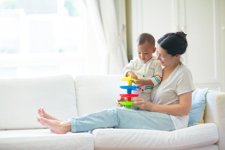 Mother and child on a white couch. Asian mom and baby boy playing in sunny bedroom. Parent and little kid relaxing at home. Family having fun together. Bedding and textile for infant nurseryの写真素材