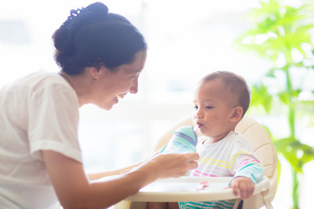 Baby eating in high chair. Asian baby boy with drink and lunch. Infant weaning and introducing solid foods. Kid in white sunny kitchen. Healthy nutrition for little child. Kids eat.の写真素材