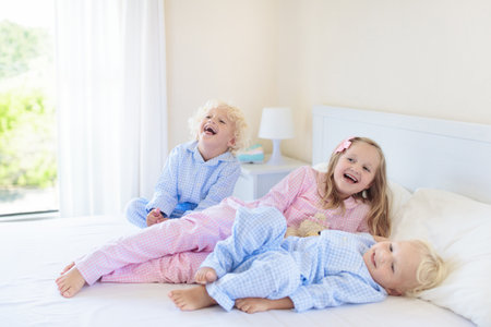 Kids playing in parents bed. Children wake up in sunny white bedroom. Boy and girl play in matching pajamas. Sleepwear and bedding for child and baby. Nursery interior for toddler kid. Family morning.の写真素材