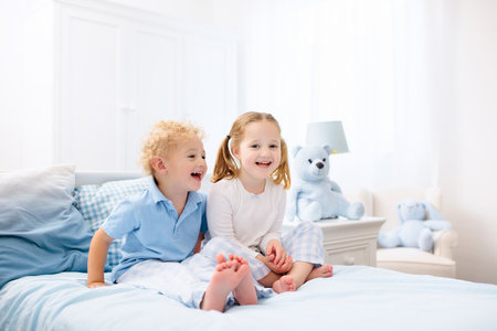 Happy kids playing in white bedroom. Little boy and girl, brother and sister play on the bed wearing pajamas. Nursery interior for children. Nightwear and bedding for baby and toddler. Family at home.の写真素材