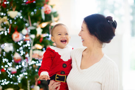 Asian family with kids at Christmas tree and fireplace. Mother and children opening gifts at fire place. Baby and mom open presents. Winter holidays interior decoration. Child in pajamas on Xmas Eve.の写真素材
