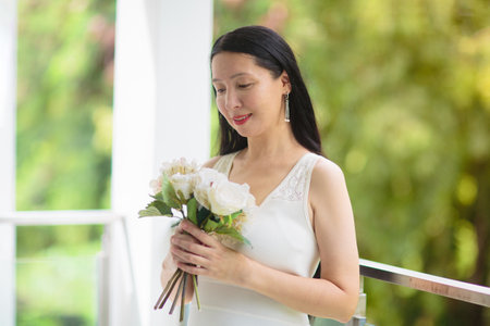 Asian woman in white dress with flower bouquet. Young bride getting ready for party. Beautiful female with peony flowers. Wedding or birthday celebration.の写真素材