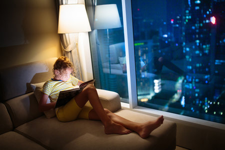 Child reading in dark room. Kids read book. Little boy reading a story on a couch at home at a big window with night city view. Cozy evening at home in modern high rise building.の写真素材