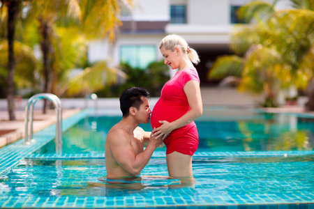 Pregnant woman in swimming pool on tropical beach holiday. Interracial couple expecting baby....