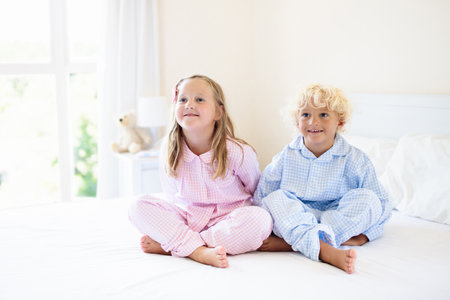 Kids playing in parents bed. Children wake up in sunny white bedroom. Boy and girl play in matching pajamas. Sleepwear and bedding for child and baby. Nursery interior for toddler kid. Family morning.の写真素材