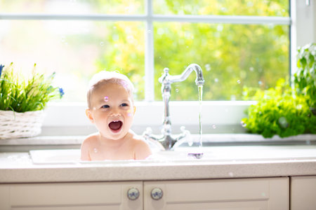 Baby taking bath in kitchen sink. Child playing with foam and soap bubbles in sunny bathroom with window. Little boy bathing. Water fun for kids. Hygiene and skin care for children. Bath room interiorの写真素材
