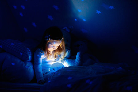 Little girl reading a book in bed. Dark bedroom with night light projecting stars on room ceiling. Kids nursery and bedding. Children read before bedtime. Toddler child playing with lamp and bear toy.の写真素材