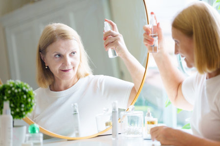 Skin care for senior woman. Anti-aging cream. Beautiful elderly lady applying anti wrinkle lifting serum in sunny bedroom with big mirror. Beauty and cosmetics product.の写真素材