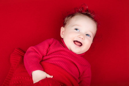 Cute baby on red knitted blanket. Winter clothing for kids. Newborn child relaxing in bed. Nursery for young children. Textile and bedding. Family morning at home. Happy little girl in wool sweater.の写真素材