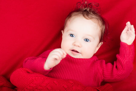 Cute baby on red knitted blanket. Winter clothing for kids. Newborn child relaxing in bed. Nursery for young children. Textile and bedding. Family morning at home. Happy little girl in wool sweater.の写真素材