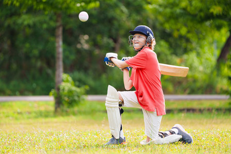 Kids playing cricket in summer park. Boy with bat and ball on cricket pitch. Helmet and guard for...