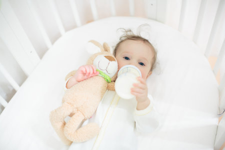 Adorable baby in white sunny bedroom. Newborn child relaxing in bed. Nursery for young children. Textile and bedding for kids. Family morning at home. New born kid during tummy time with toy bear.の写真素材
