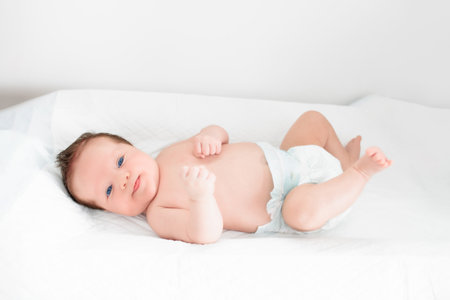 Baby in diaper in white sunny bedroom. Newborn child relaxing in bed. Nursery for young children. Textile and bedding for kids. Family morning at home. New born kid in nappy.の写真素材
