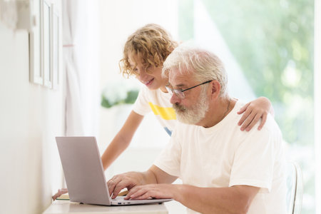 Senior man working on computer with grandchild. Grandfather and child having video chat or call with family. Online shopping  and social media in retirement. Retired elderly male with laptop at home.の写真素材