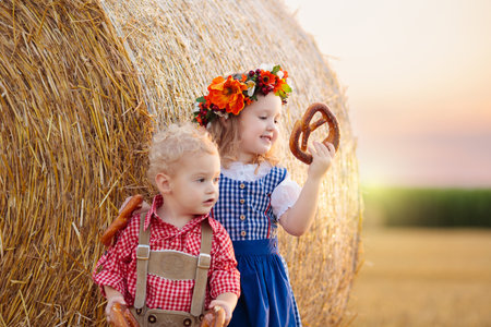 Child in wheat field with bread and pretzel. Oktoberfest time in Germany. Kids in traditional German dirndl dress and leather pants in autumn harvest crop field.の写真素材