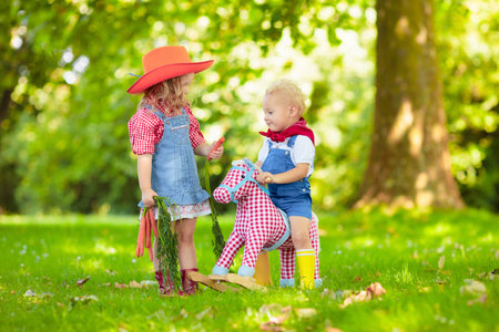 Little boy and girl dressed up as cowboy and cowgirl playing with toy rocking horse in park. Kids play outdoors. Children in Halloween costumes at trick or treat. Toys for preschooler or toddler childの写真素材