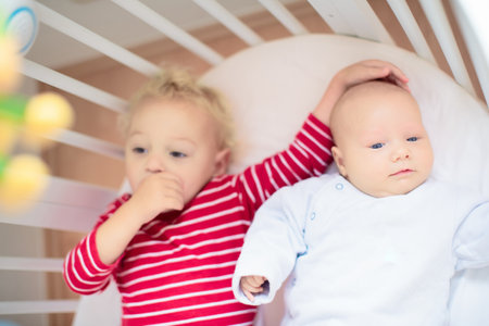 Child watching newborn baby in bassinet. Sibling meeting new family member. Kids play in white bedroom. Nursery crib bedding and textile for young kid. Siblings love.の写真素材