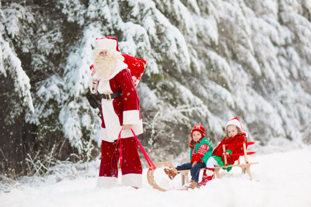 Kids and Santa with Christmas gifts in snowy forest. Children help Santa Claus in snowy park. Fun sleigh ride. Xmas vacation fun. Santa with presents and elves. Winter season holiday.の写真素材