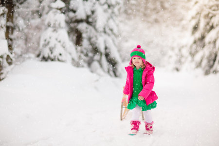 Children playing with snow in winter. Little girl in colorful jacket and knitted hat riding sleigh in winter park on Christmas. Kids sled in snowy forest. Sledge riding for children.の写真素材
