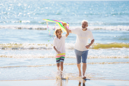 Grandfather and kids play on summer beach. Family day outdoor. Grandparent and grandchildren fly kite, travel to sea side. Generation love. Active senior man playing with grandson. Happy retirement.の写真素材