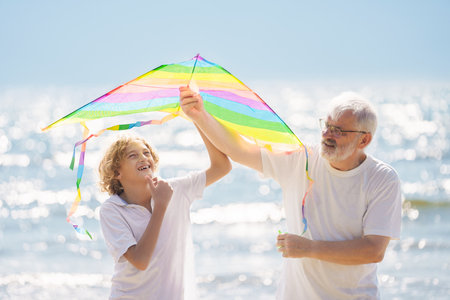 Grandfather and kids play on summer beach. Family day outdoor. Grandparent and grandchildren fly kite, travel to sea side. Generation love. Active senior man playing with grandson. Happy retirement.の写真素材