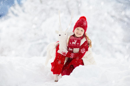 Child playing with reindeer in snow on Christmas vacation. Winter outdoor fun. Kids play in snowy park on Xmas Eve. Little girl in knitted sweater, hat, scarf and mittens with Christmas decoration.の写真素材