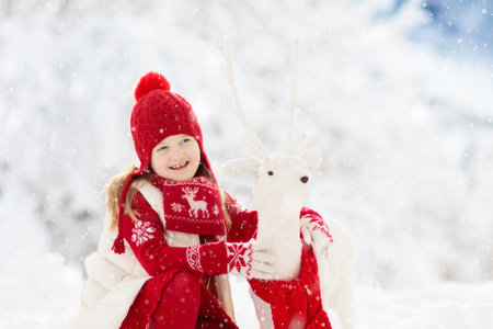 Child playing with reindeer in snow on Christmas vacation. Winter outdoor fun. Kids play in snowy park on Xmas Eve. Little girl in knitted sweater, hat, scarf and mittens with Christmas decoration.の写真素材