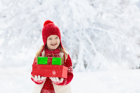 Child in red hat with Christmas presents and gifts in snow. Winter outdoor fun. Kids play in snowy park on Xmas Eve. Little girl in knitted sweater, scarf and mittens with Christmas decoration.の写真素材
